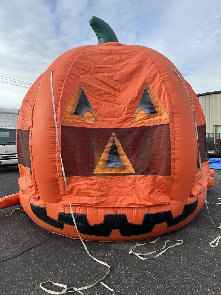 A massive, bright orange inflatable pumpkin bounce house with a green stem and a smiling jack-o'-lantern face, set up for a fall festival in a grassy field in Ohio.