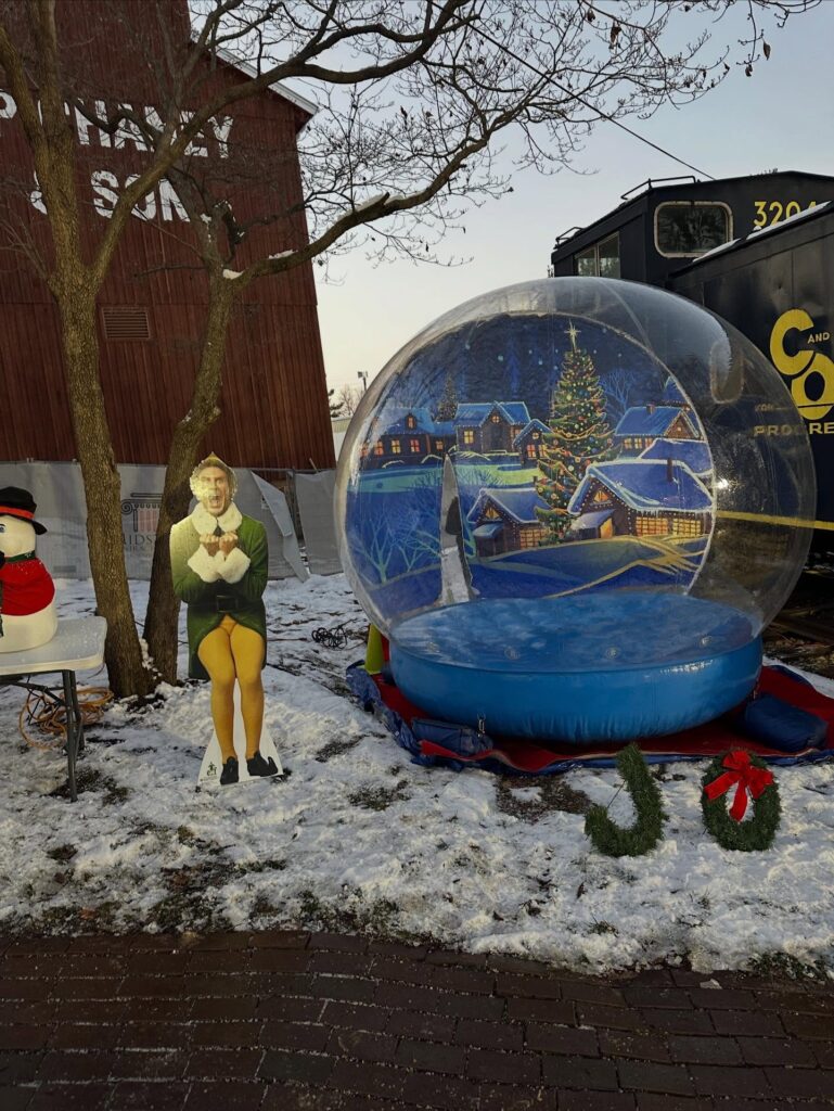 A large, transparent inflatable snow globe with a festive winter backdrop, filled with artificial snow and happy guests posing for a holiday photo at a Central Ohio event.